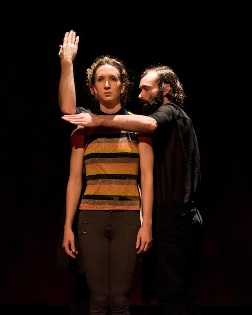 A male dancer stands next to a female dancer. His arms surround her shoulder making a ninety degree angle. The female dancer dressed in a striped tank stares out to the audience